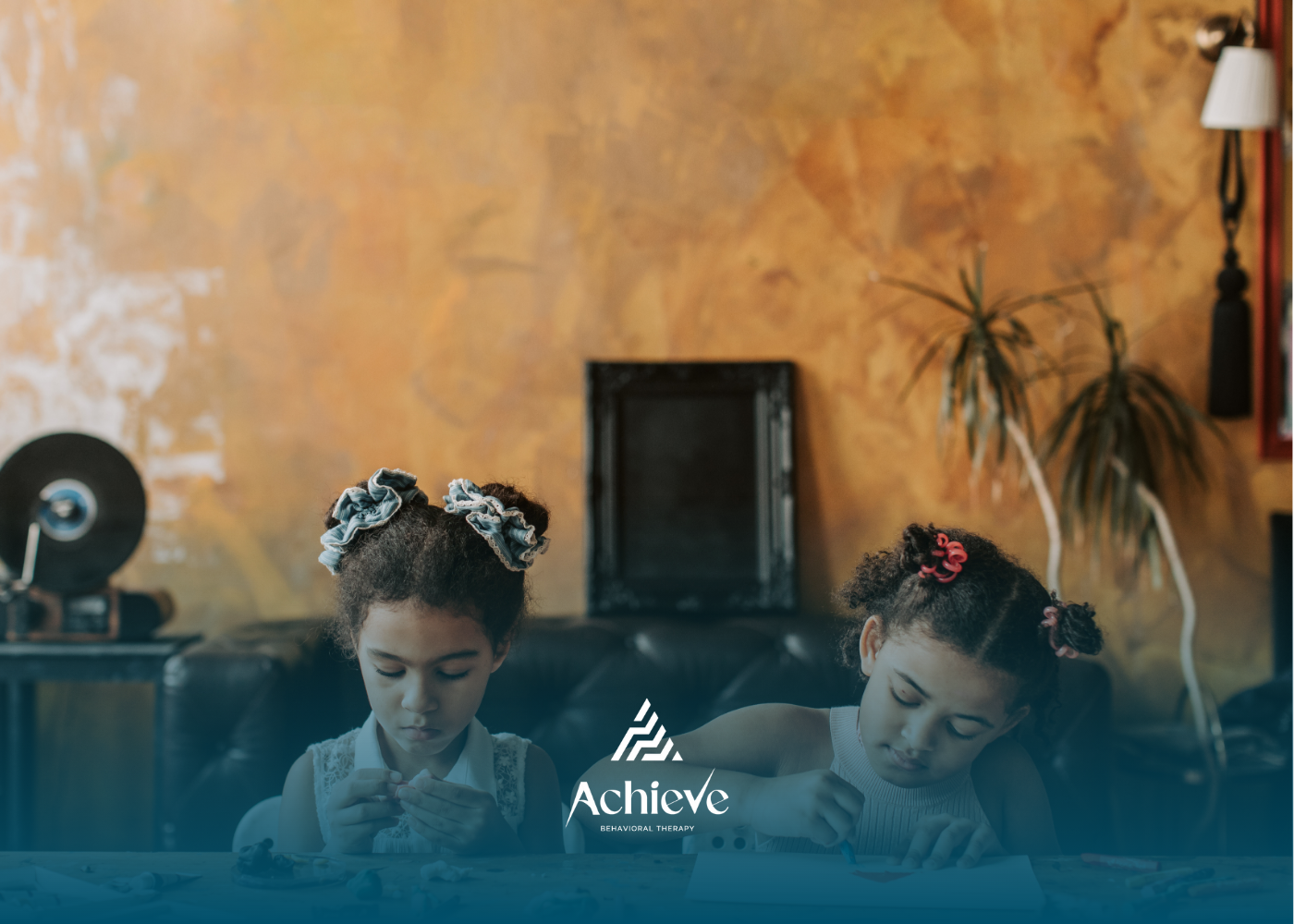 Two girls with bows in their hair sit, looking down at something on a table. In front of a decorated wall.