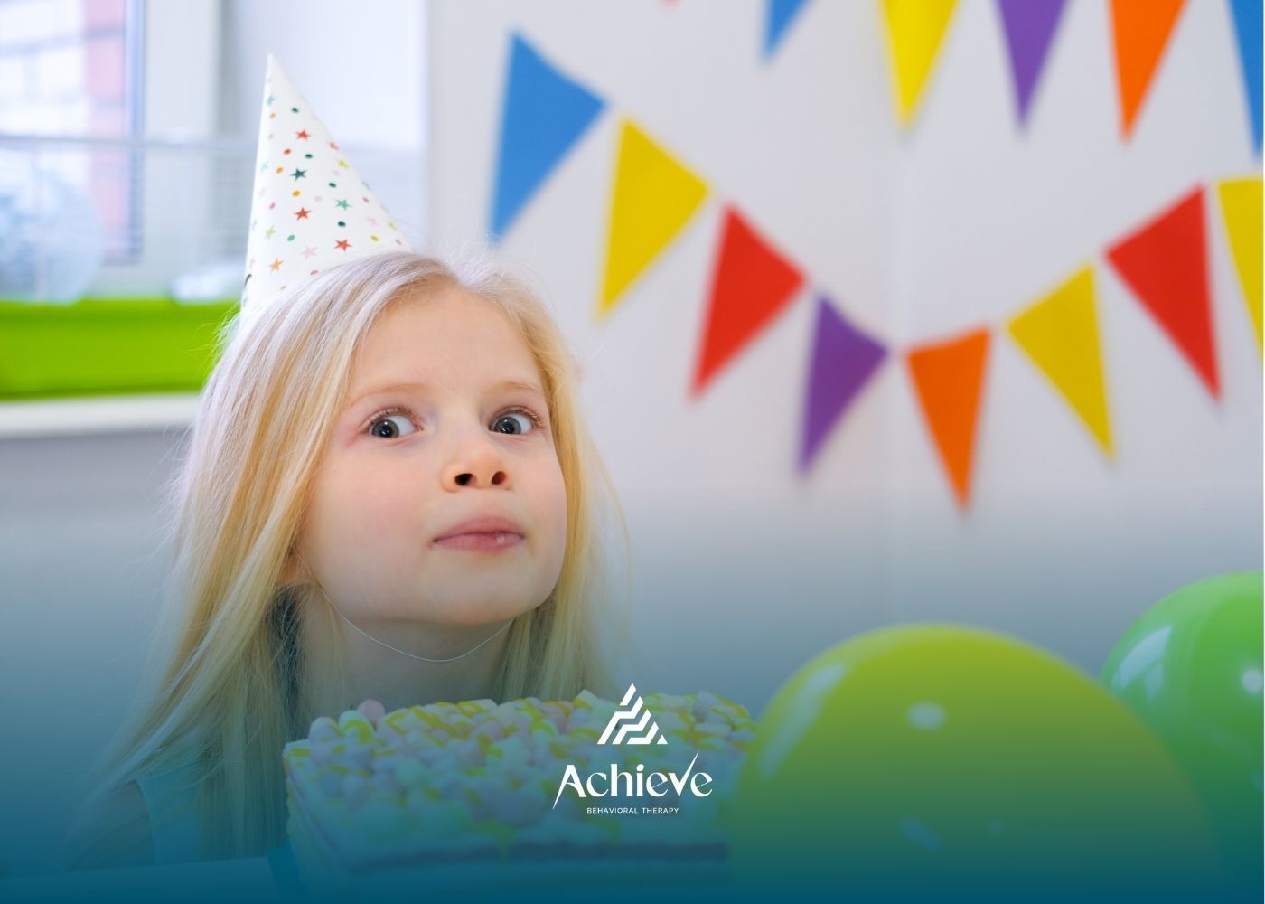 Girl with party hat looking at cake, colorful flags in the background, green balloons.