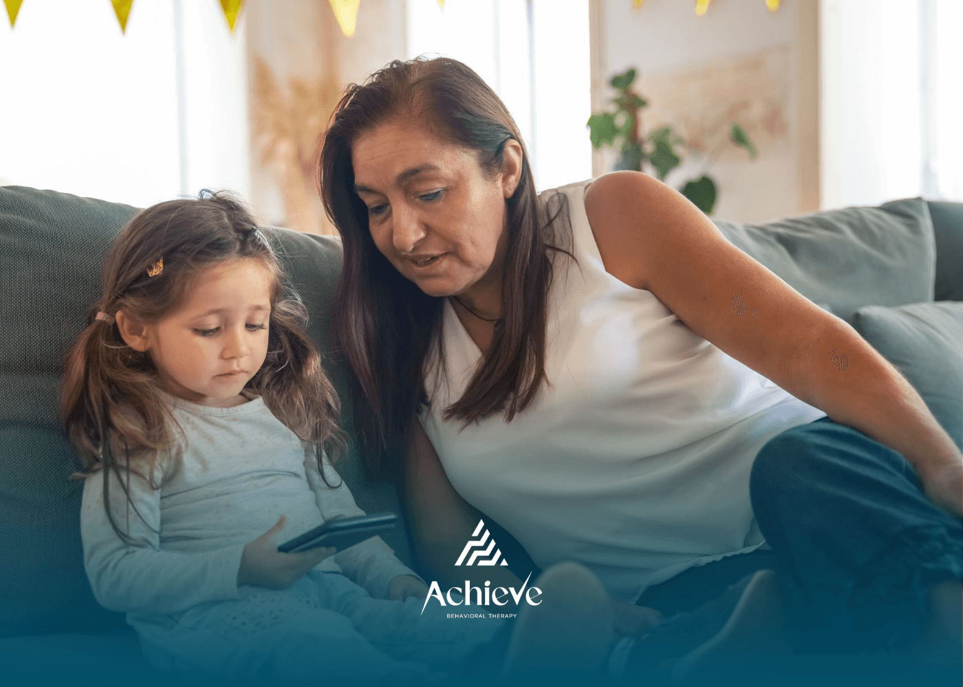 RBT and young autistic girl sit on couch, focused on phone screen during ABA therapy in New Jersey.