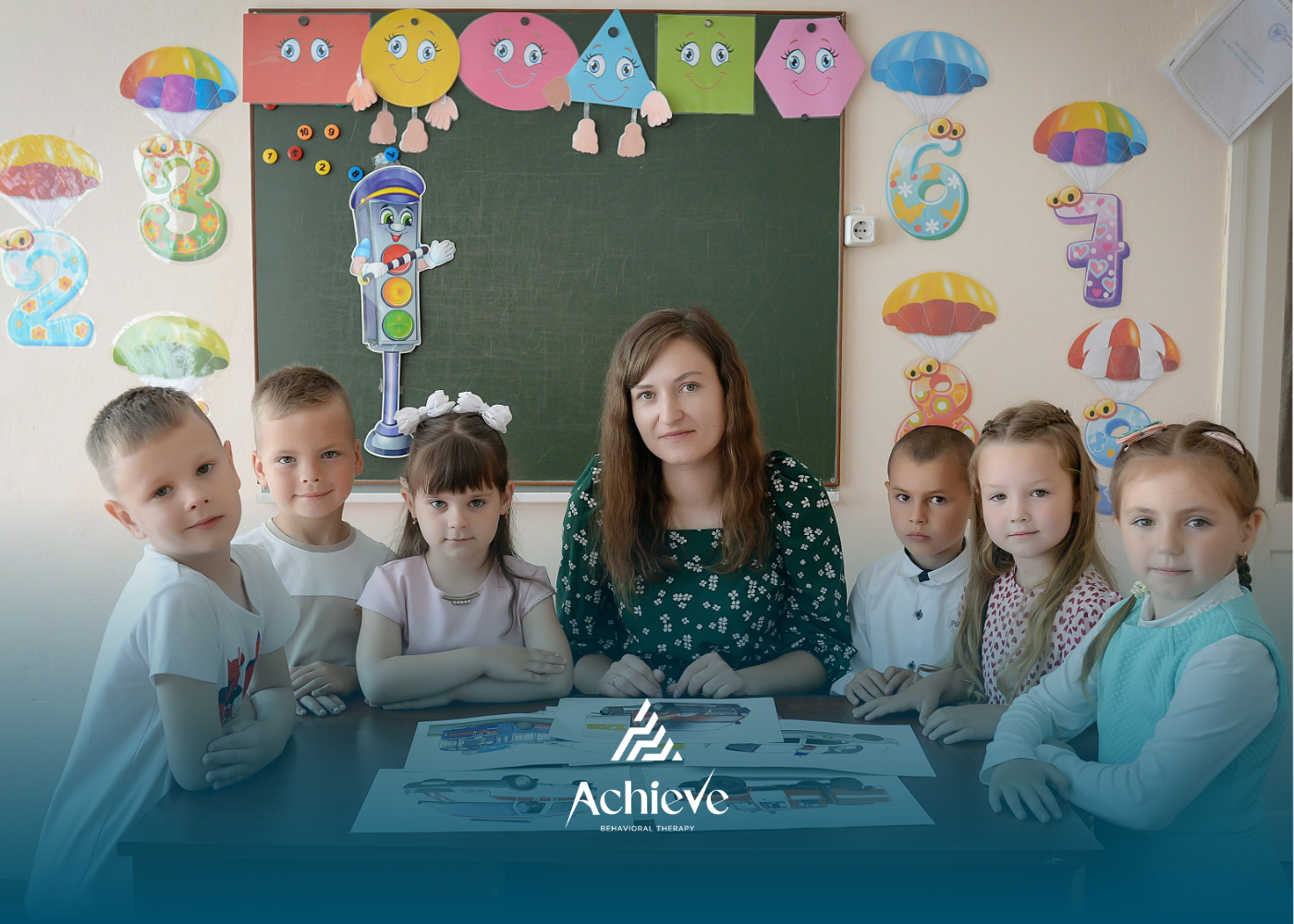 A teacher and six children sit at a table in a classroom with educational wall decorations and a chalkboard background.