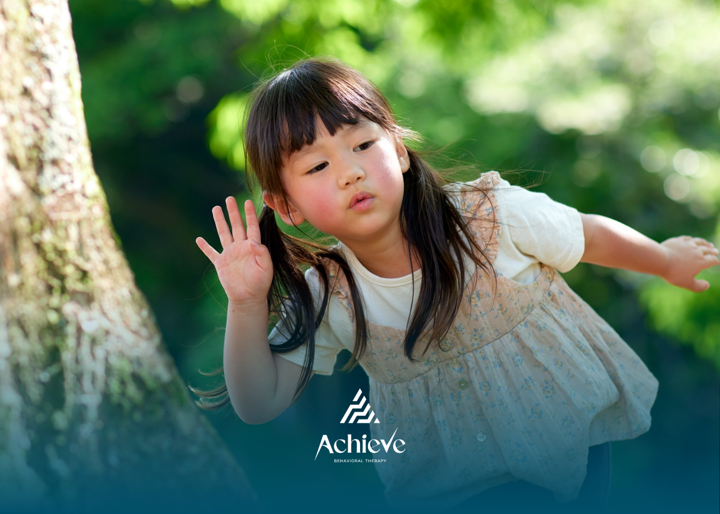 Child leaning near a tree in a sunlit green park, hand raised as if waving, wearing a light dress.