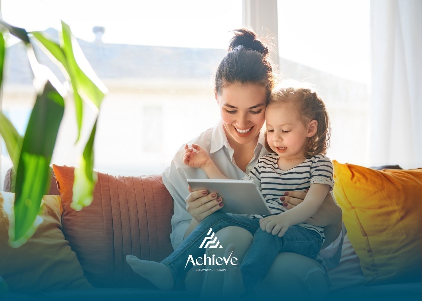 Woman and child looking at a tablet, smiling on a couch indoors, near a window.