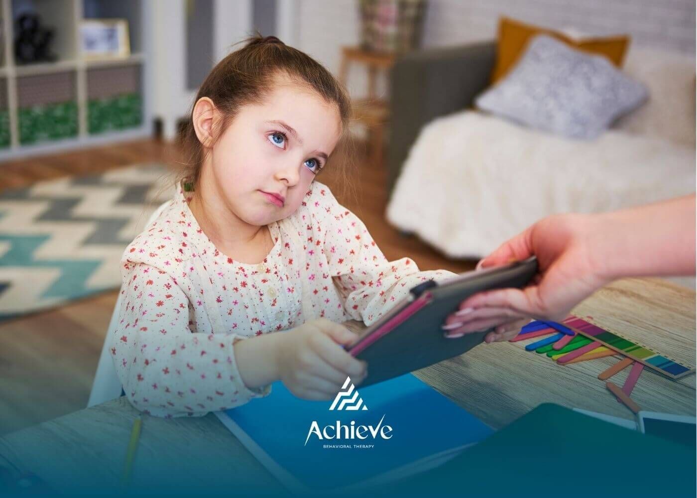 Autistic girl looking disinterested while holding a tablet, as an adult helps her with her work.