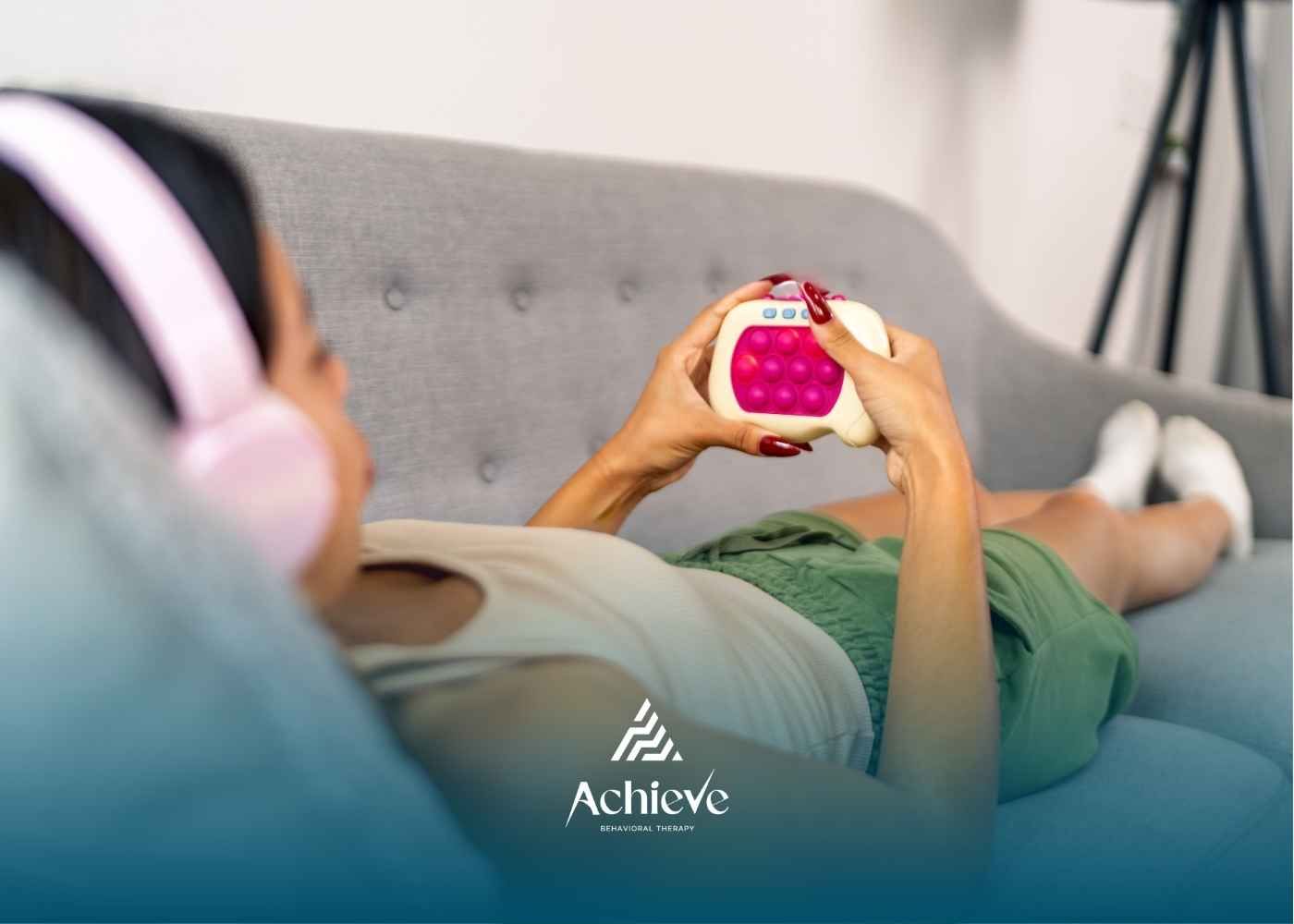 A person lies on a grey sofa wearing pink headphones, holding a fidget toy with pink buttons.