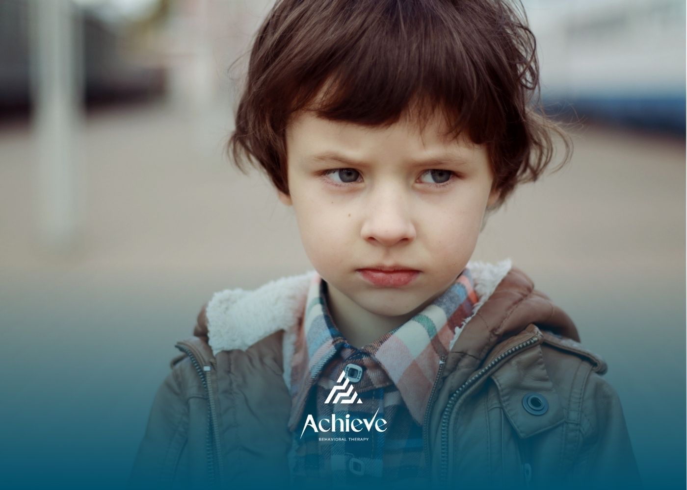 A young child with a sullen expression wears a jacket and looks off to the side; outdoor setting.