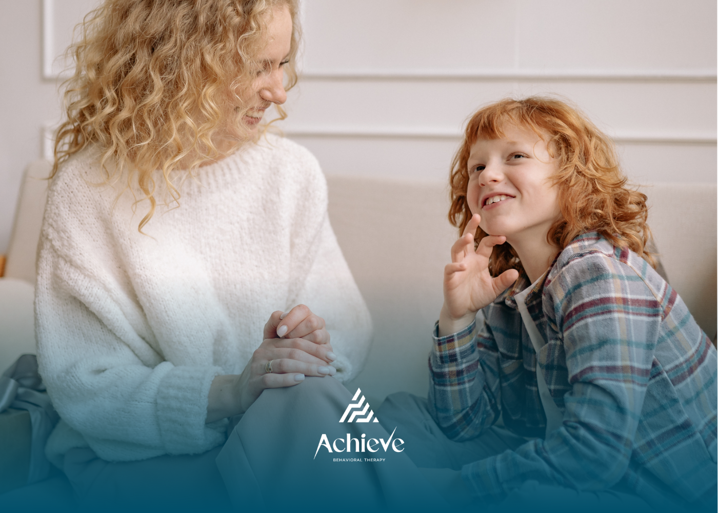 A person with wavy blonde hair and a person with curly red hair smiling and talking, sitting together in a bright room.