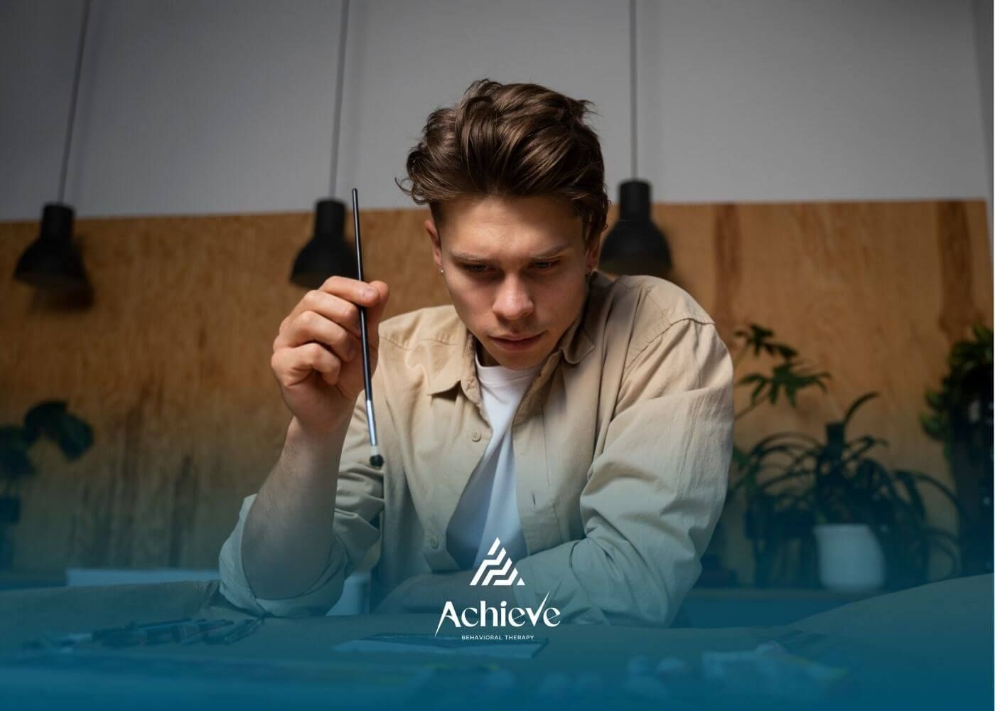 A young man concentrating while holding a paintbrush, preparing to create artwork on a table.