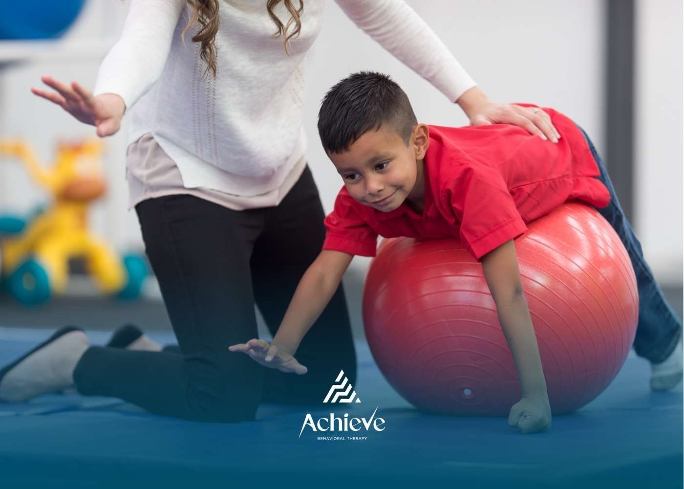 Boy on a red exercise ball being assisted by an adult, in a therapy setting.