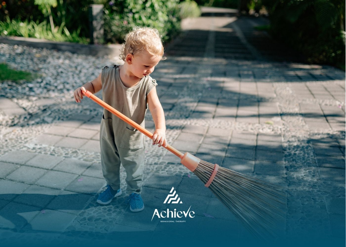 Child sweeping a paved walkway with a broom; sunny outdoor setting.