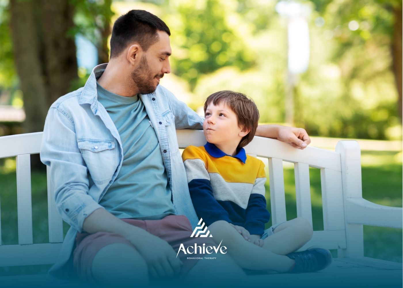 Man and boy sitting on a white bench, looking at each other in a park.