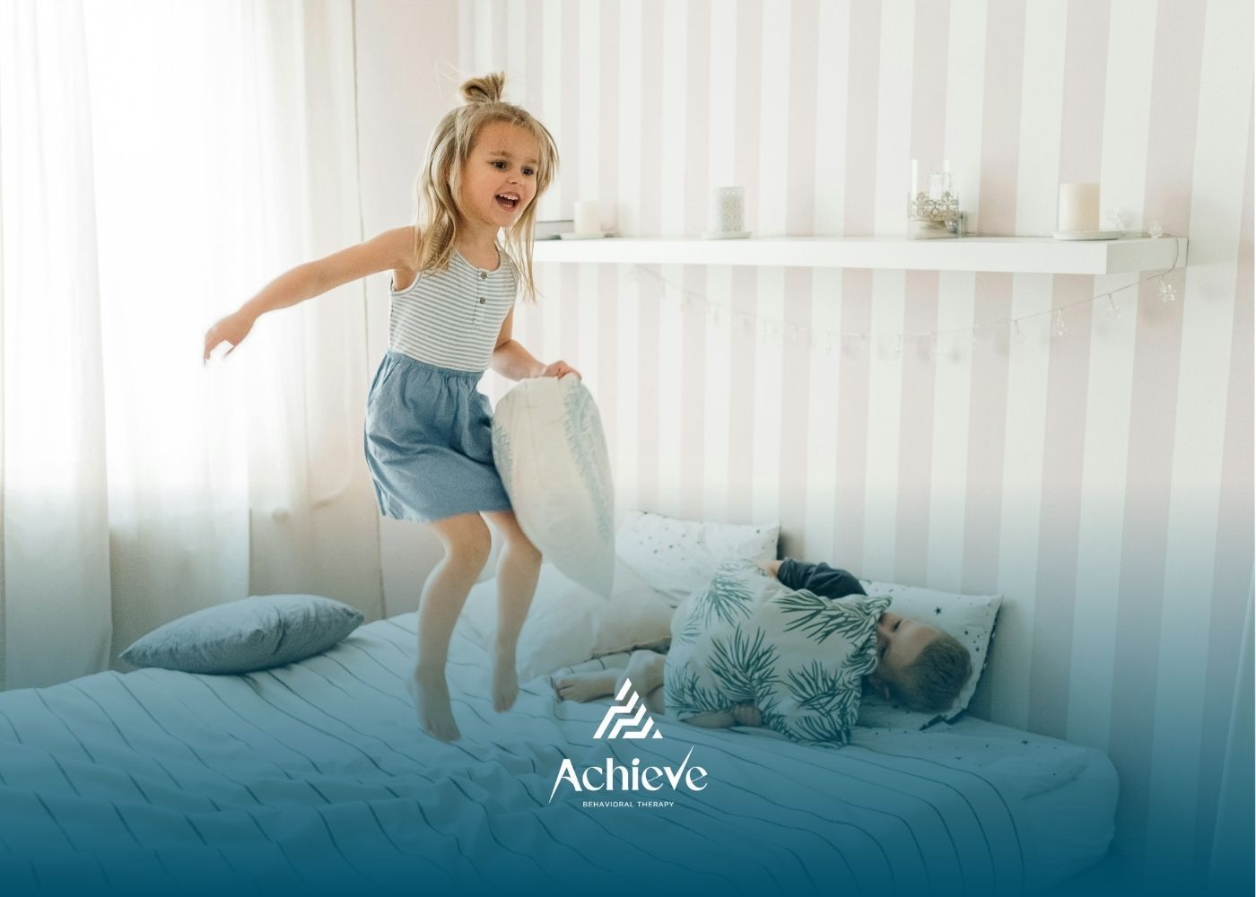 Girl jumping on bed, holding a pillow, with another person lying down. Bedroom setting.
