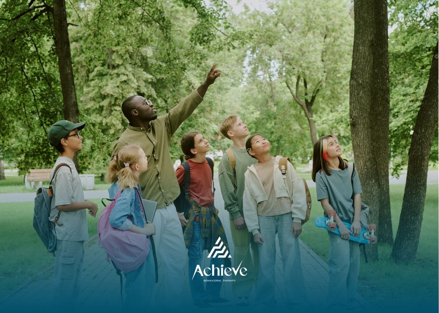 Group of children and adult looking up at trees in a park. Person points upwards.