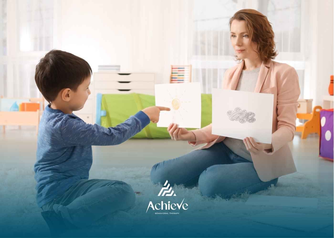 A child, seated on a rug, points to a drawing of a sun held by an adult woman in a cozy room.