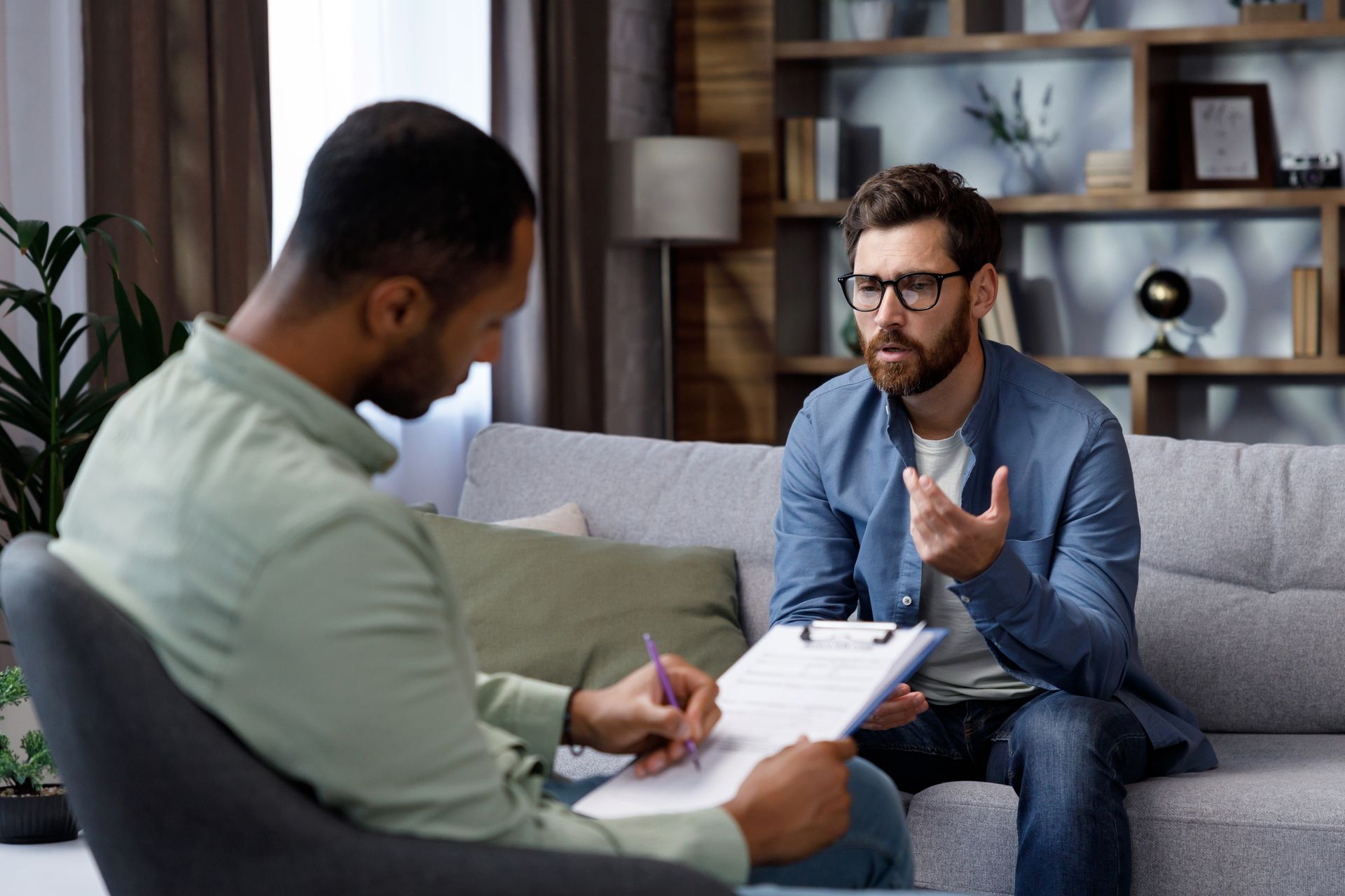 A man is sitting on a couch talking to another man while holding a clipboard.