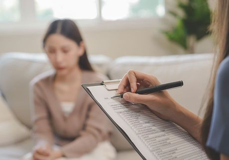 A woman is sitting on a couch while a woman writes on a clipboard.