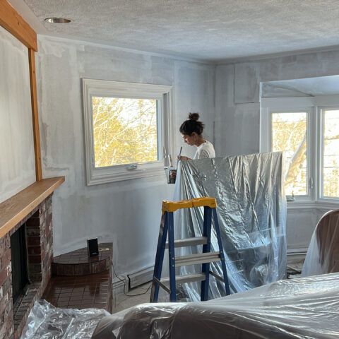 Woman painting a room, standing near a window on a ladder, covered furniture.