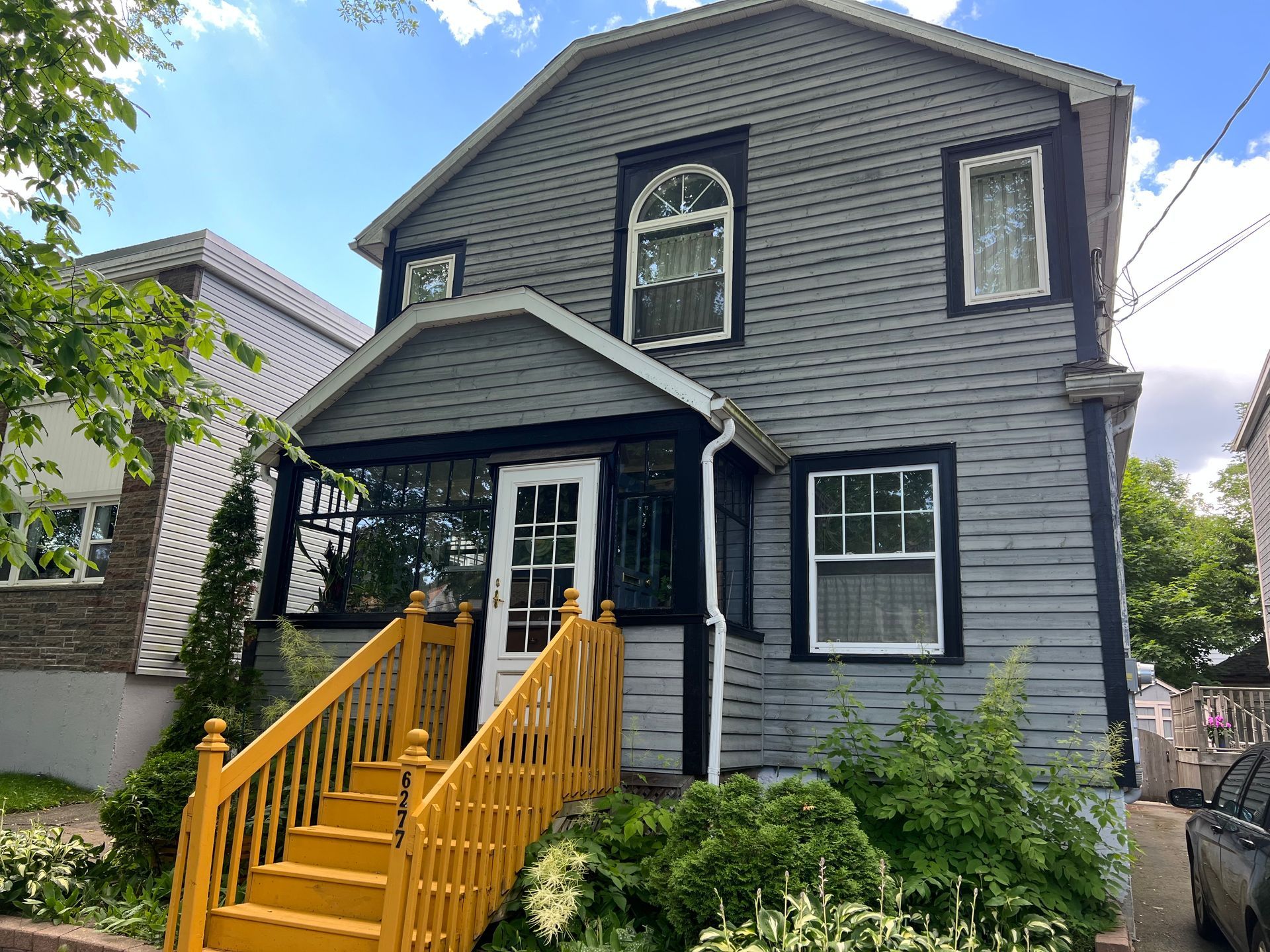 Two-story house with gray siding, yellow stairs, and white trim. Overgrown bushes in front.