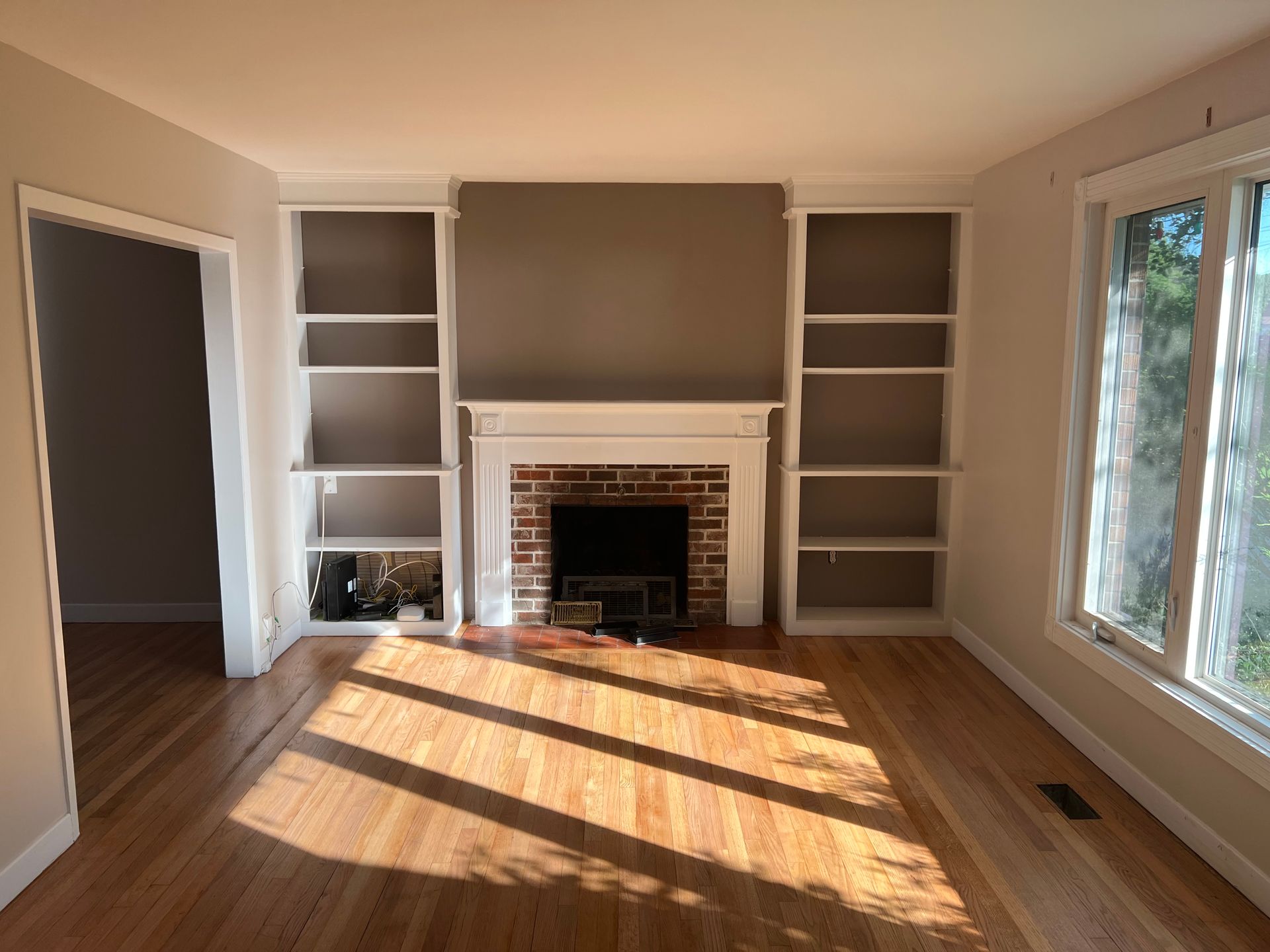 Living room with fireplace, built-in bookshelves, and wooden floor. Brown and white color scheme. Sunlight streams in.