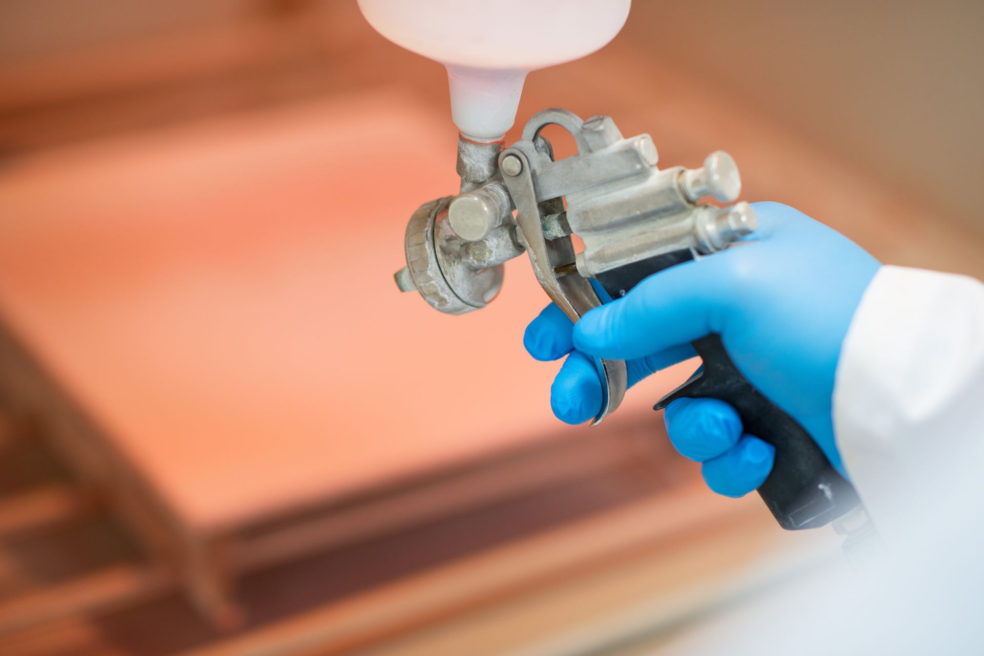 Hand with blue glove holding spray gun, applying reddish finish to wooden surface.