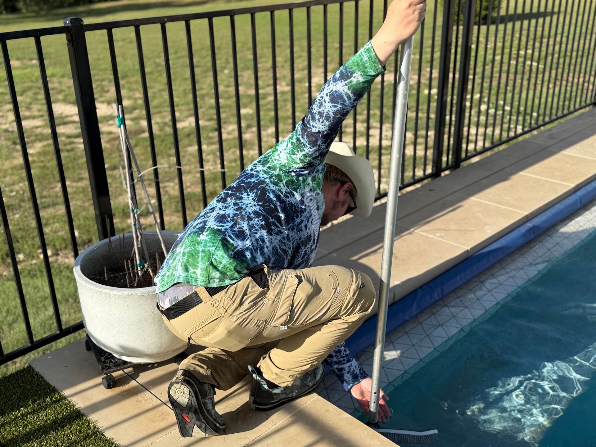 Person in tie-dye shirt checks pool water with a pole, kneeling near the pool edge.