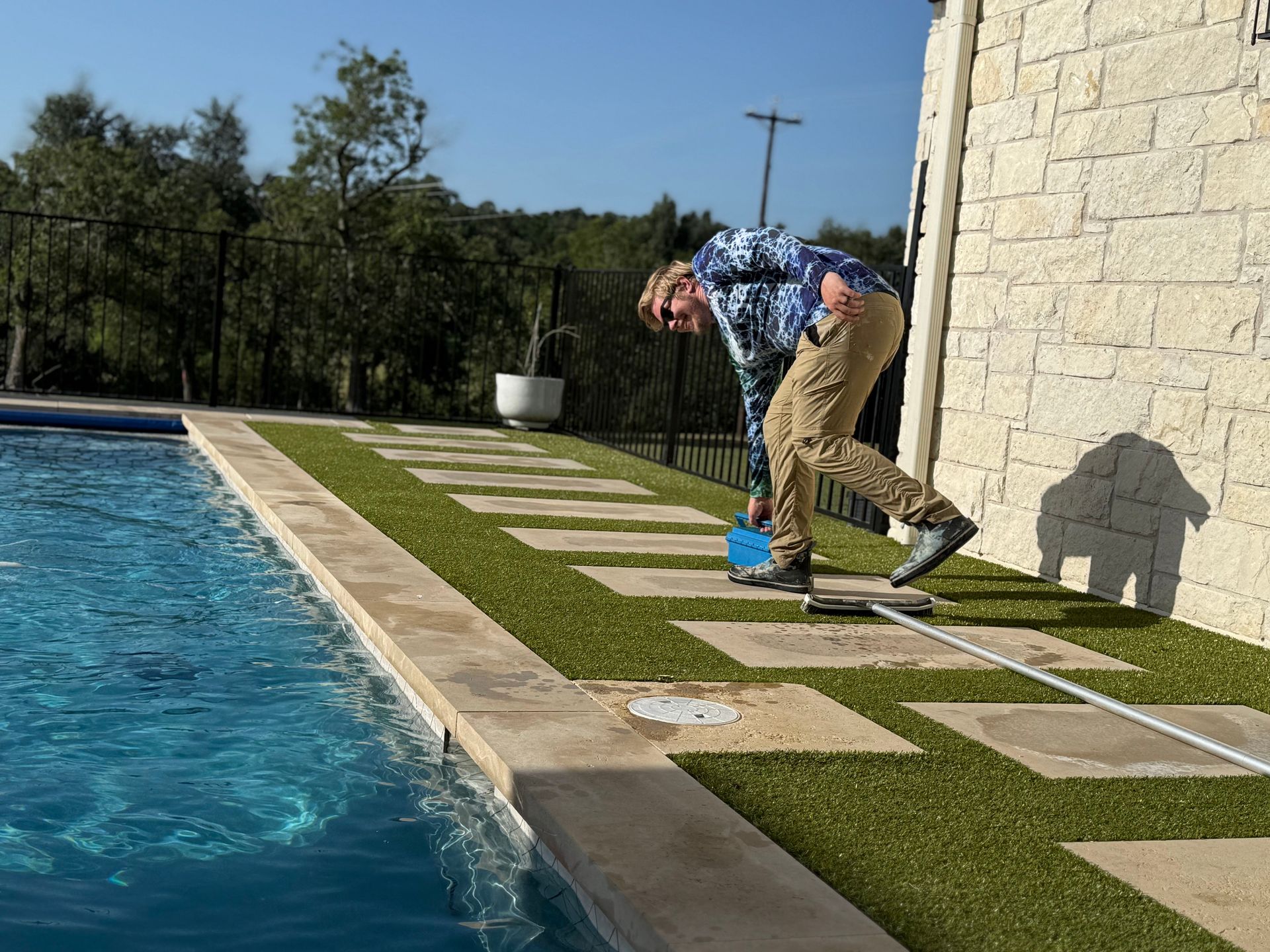 Man cleaning pool deck with a blue tool, near a pool and stone wall.