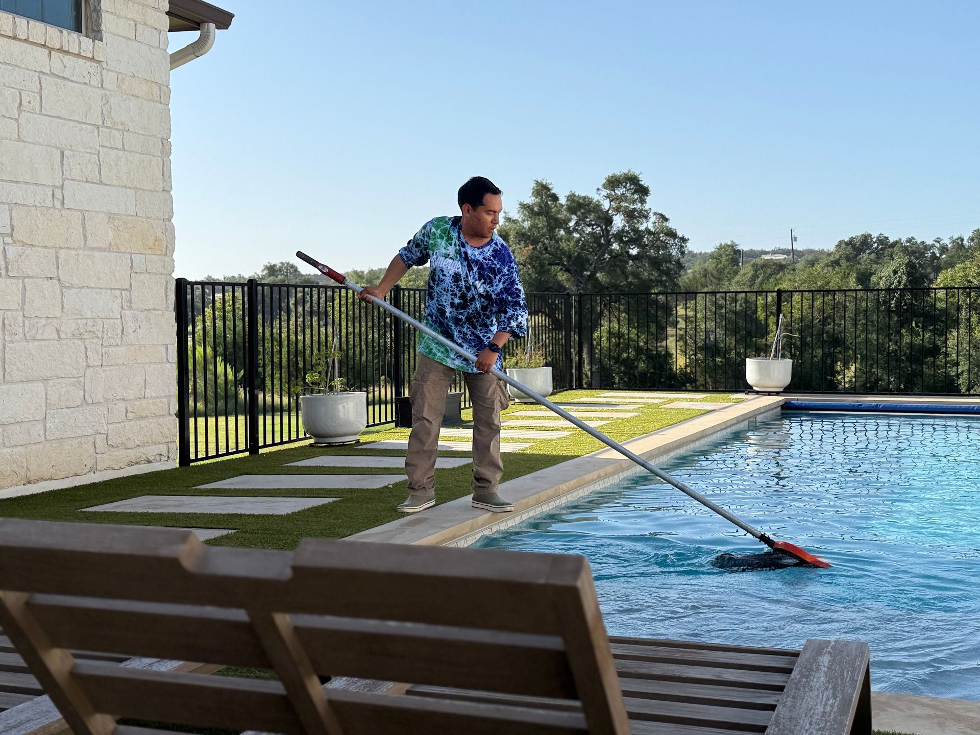 Person cleaning a pool with a long-handled net on a sunny day; backyard setting.