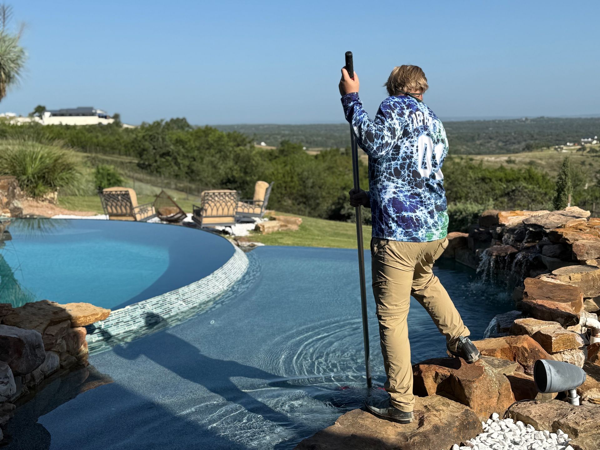 Person in blue shirt, beige pants checks a pool with a pole. Pool is near a landscape with trees, sky.