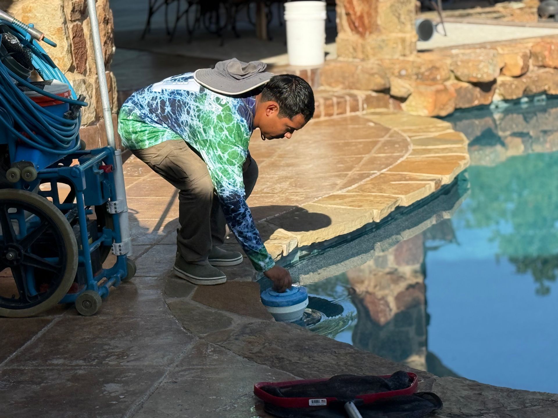 Pool cleaner adding chemicals to a pool. Man wearing a hat and colorful shirt at the edge.