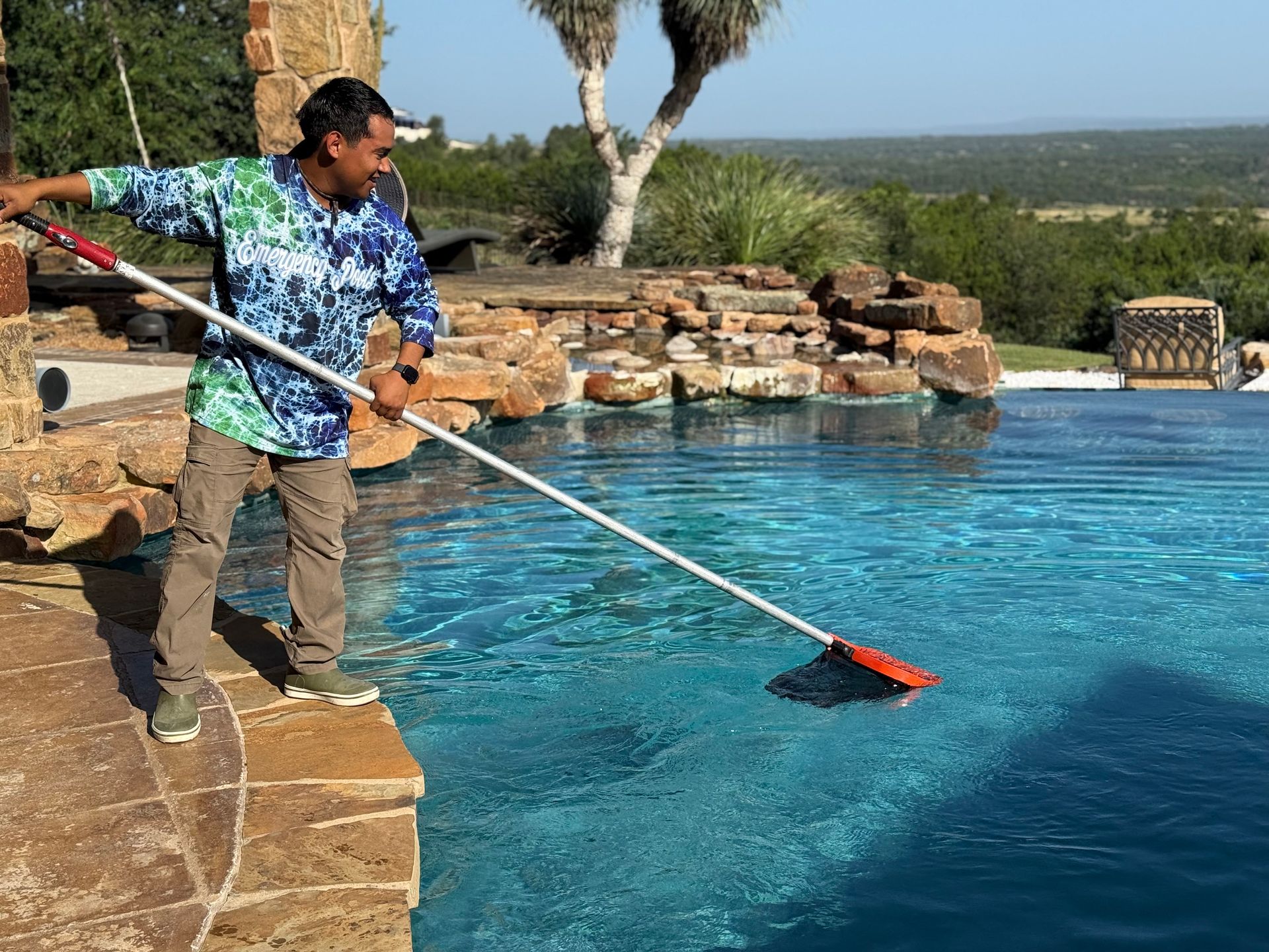 Man cleaning a turquoise pool with a long-handled brush, set against a natural landscape.