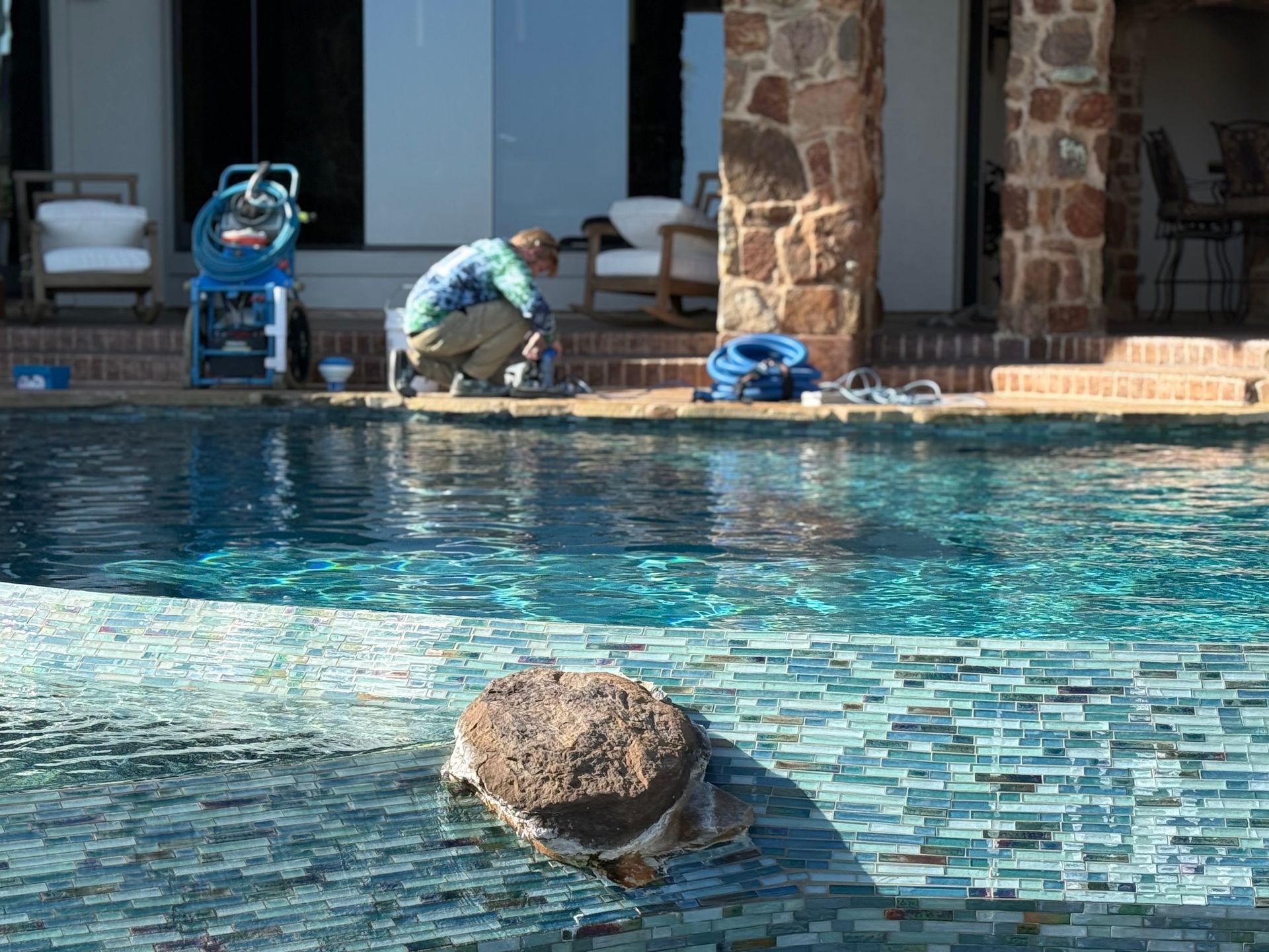Man working on pool, with hose and equipment visible. Clear blue water, stone pillar, outdoor setting.