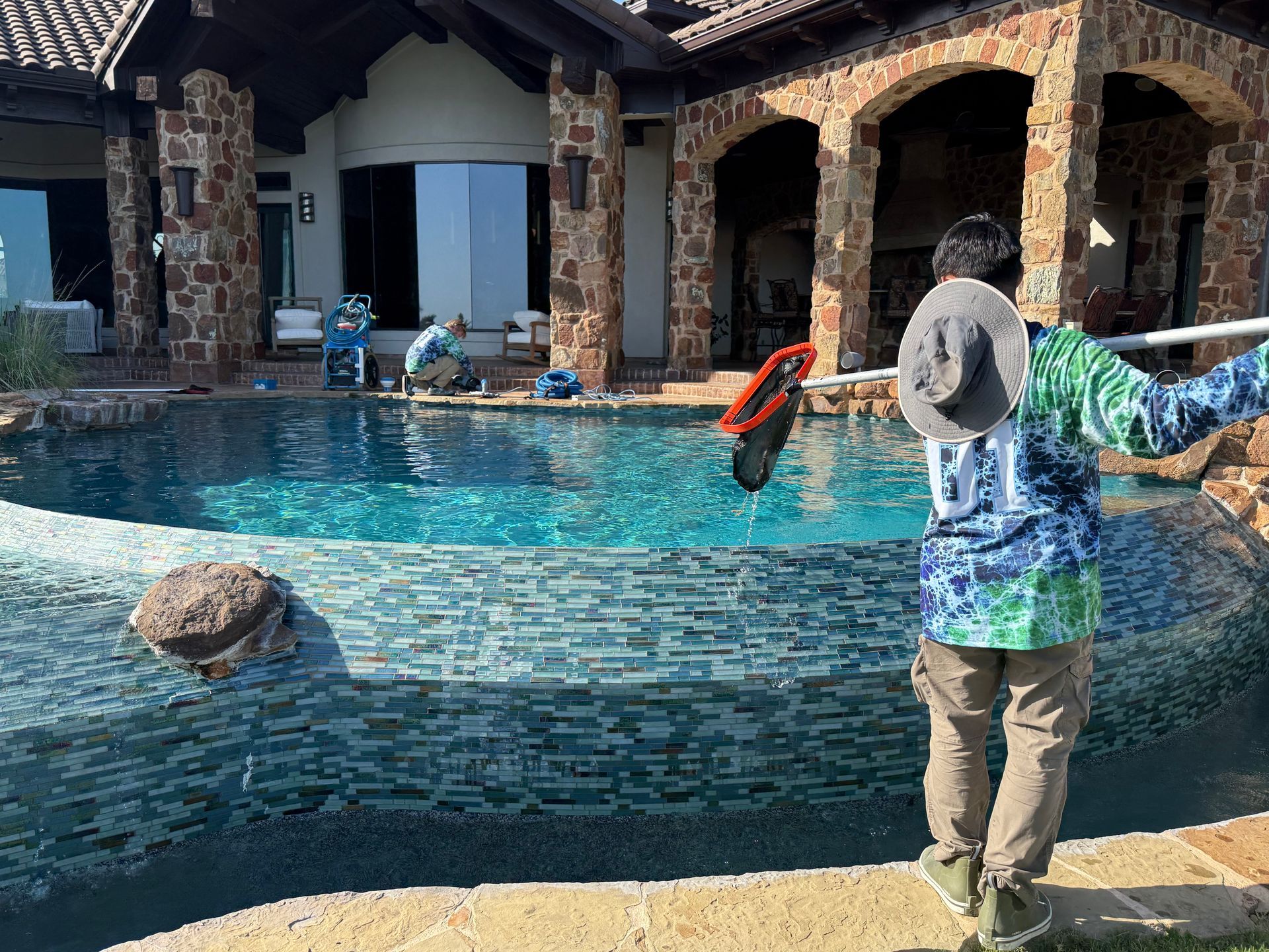 Man cleaning a luxurious pool with others in the background. Stone columns frame a house.