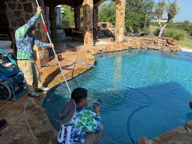 Two people cleaning a blue pool. One uses a net, the other a vacuum. Exterior, sunny day.