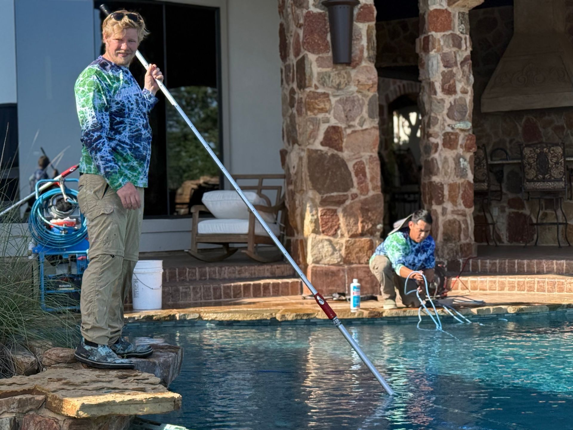 Two people cleaning a pool. One stands with a long pole, the other kneels by the water
