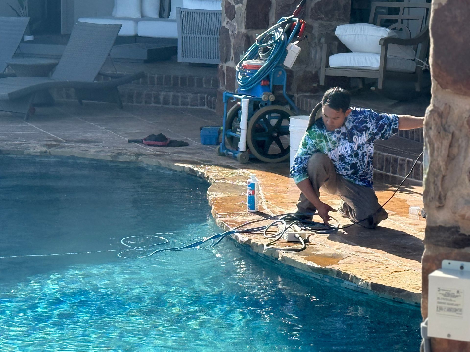 Man works on pool equipment near a pool's edge; blue water, blue/green tie-dye shirt, brown stone.