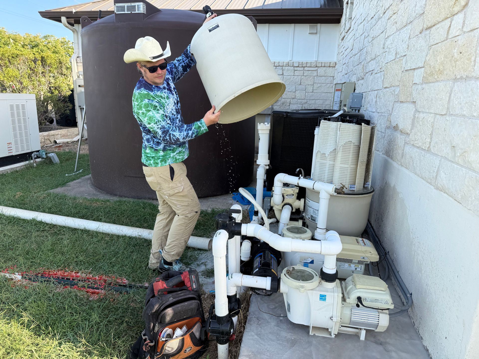 Man in cowboy hat emptying container into pool equipment near a large brown water tank outside.