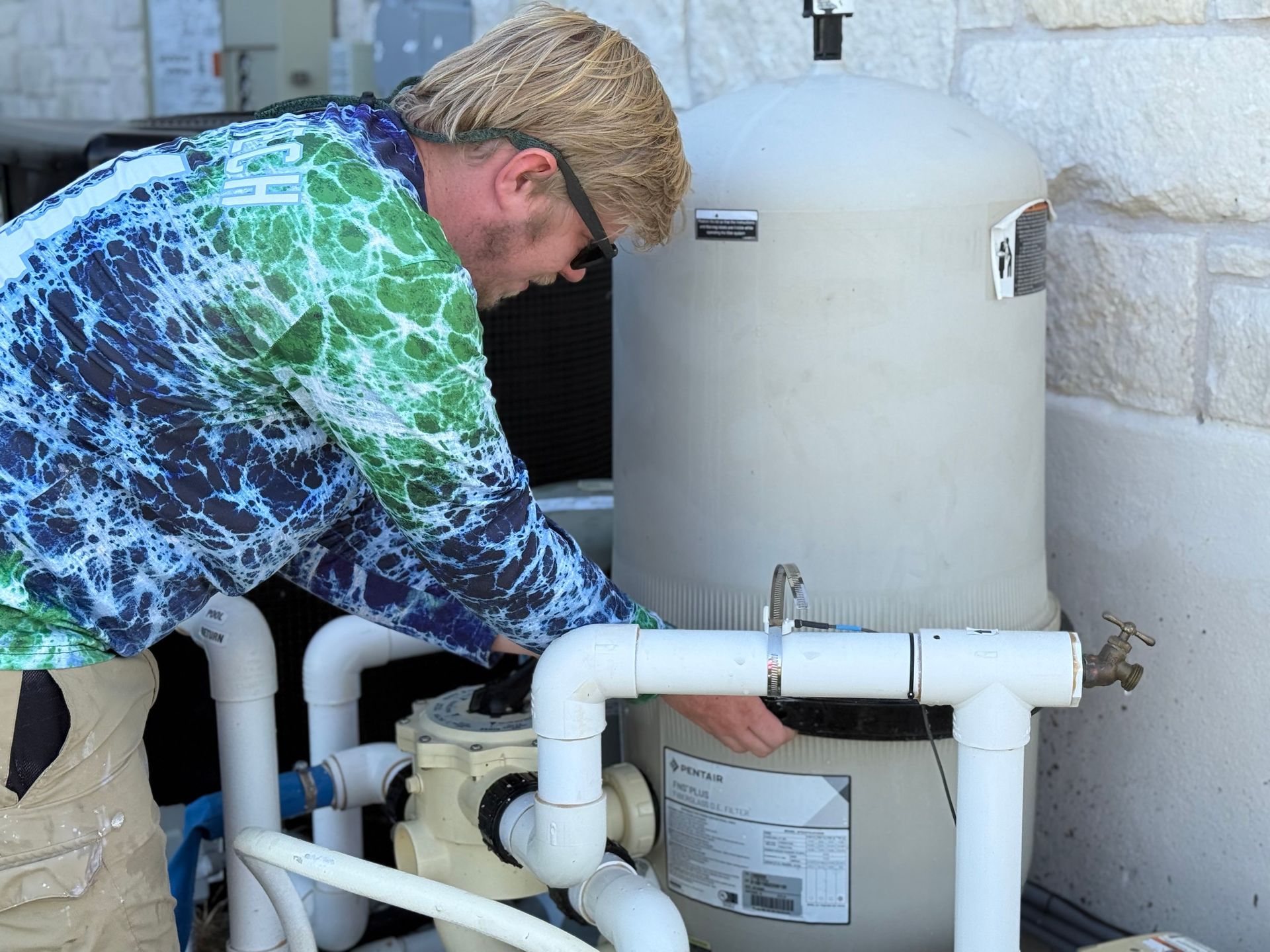 Man in tie-dye shirt adjusts pool filter; outdoors, sunny.
