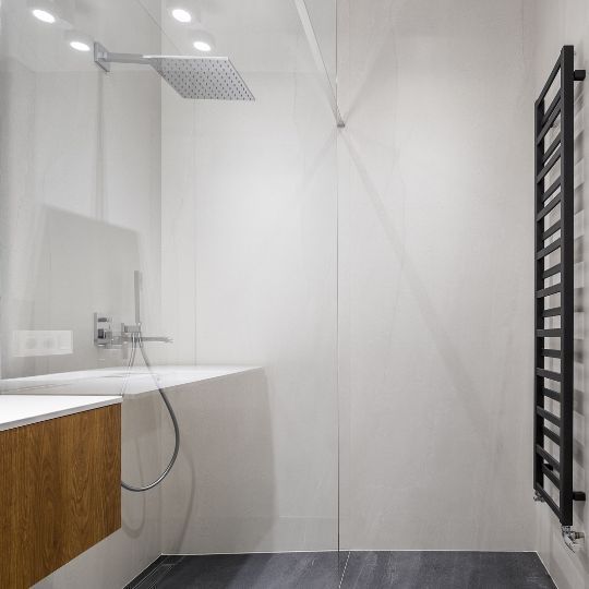 A black radiator attached to a white wall, opposite to a walk-in shower, in a modern bathroom.