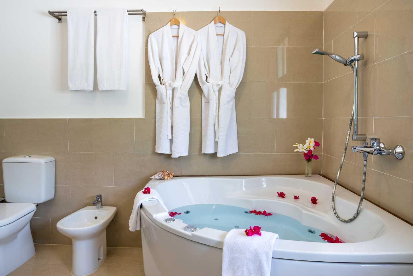 Corner bath with flowers in it, bathrobes on the wall, towels hung-up. A shower for the bath, bidet and toilet are also in the photograph.