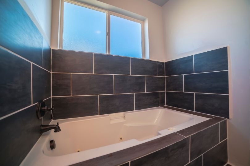 Acrylic bath in the corner of a bathroom, with grey tiles on the walls.