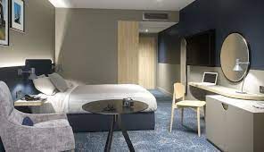 Hotel room with bed, chair, and two tables. The floor has a carpet on it, the curtains are beige.