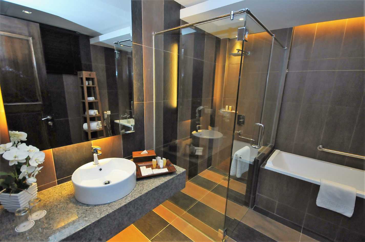 Shower with a rectangular glass panel, a white bath and a white sink. The room has a gold theme with lights coming from the perimeter of the ceiling, mirror and worktop.