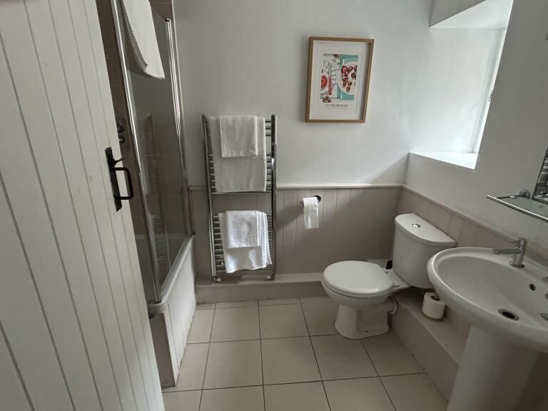 Bathroom with a bathtub, toilet, sink, and towel rack. White walls, beige tile floor. Art on the wall.