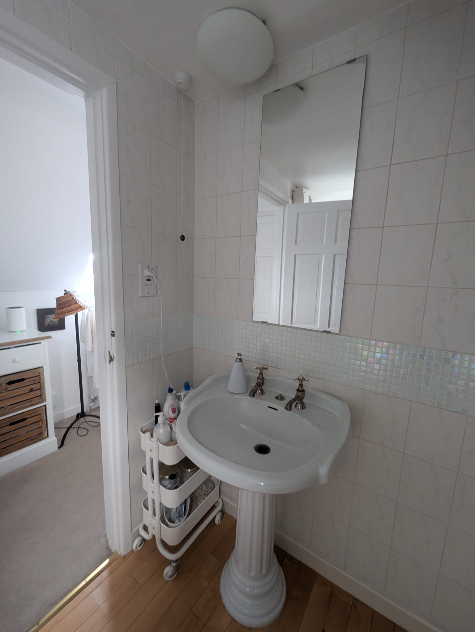 Small white bathroom with pedestal sink, tiled walls, and a rolling storage cart.