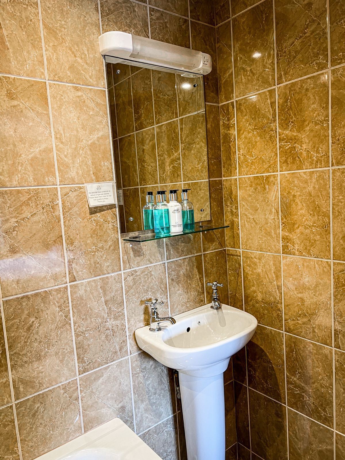 Bathroom with pedestal sink, mirror, toiletries on shelf, and tile walls.