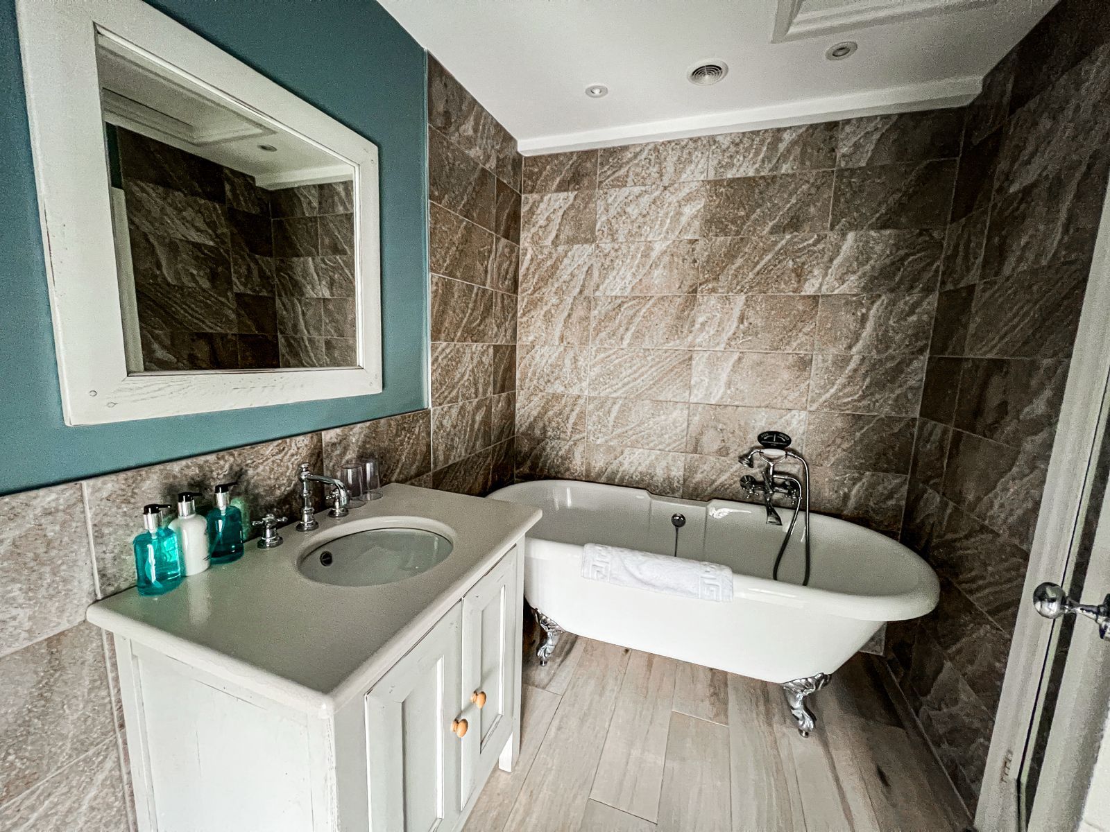 Bathroom with a white claw-foot tub, vanity, and stone tile walls. A large mirror hangs over the vanity.