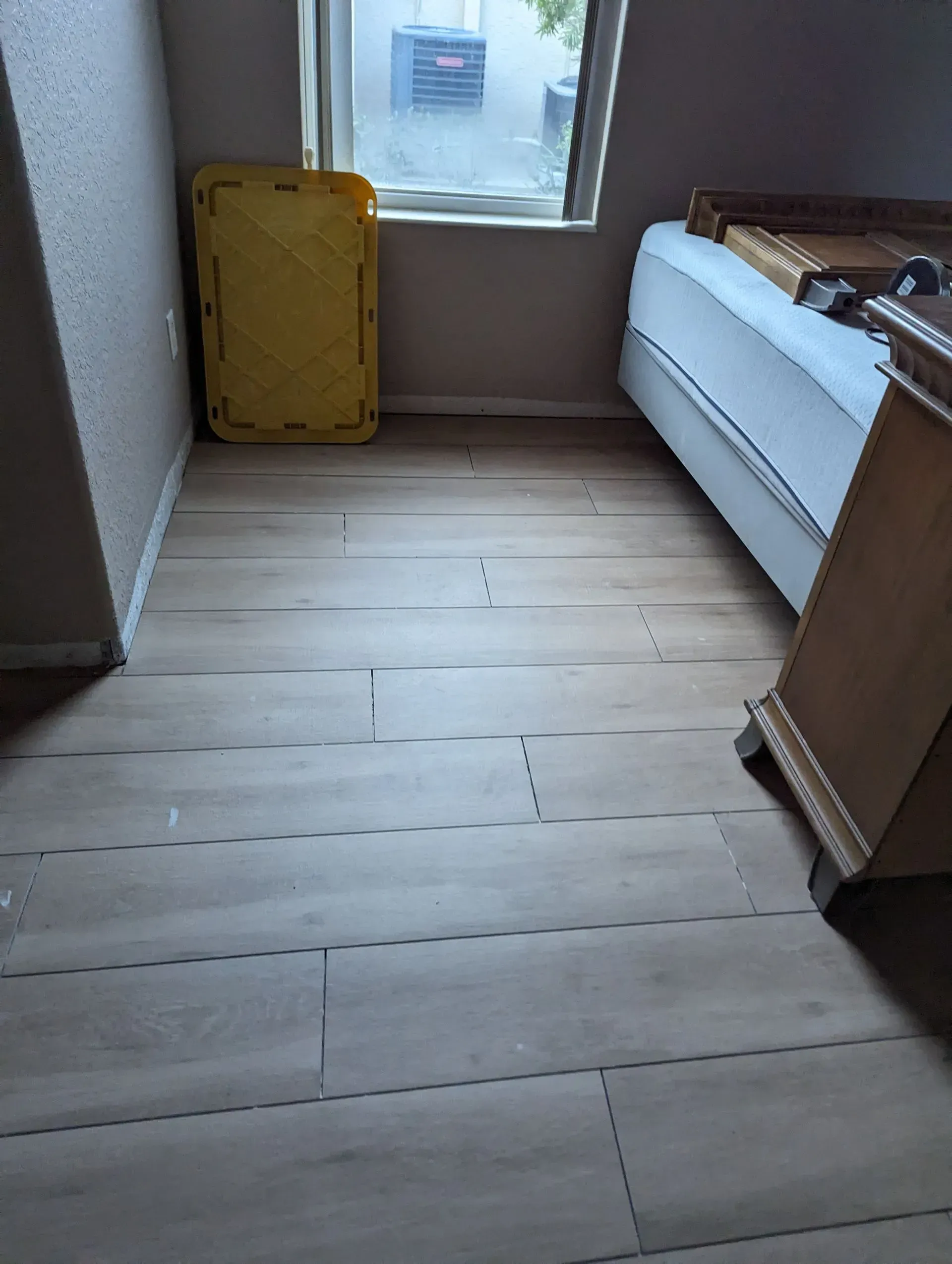 Room with light wood-look flooring, a bed, a chest of drawers, and a yellow bin near a window.