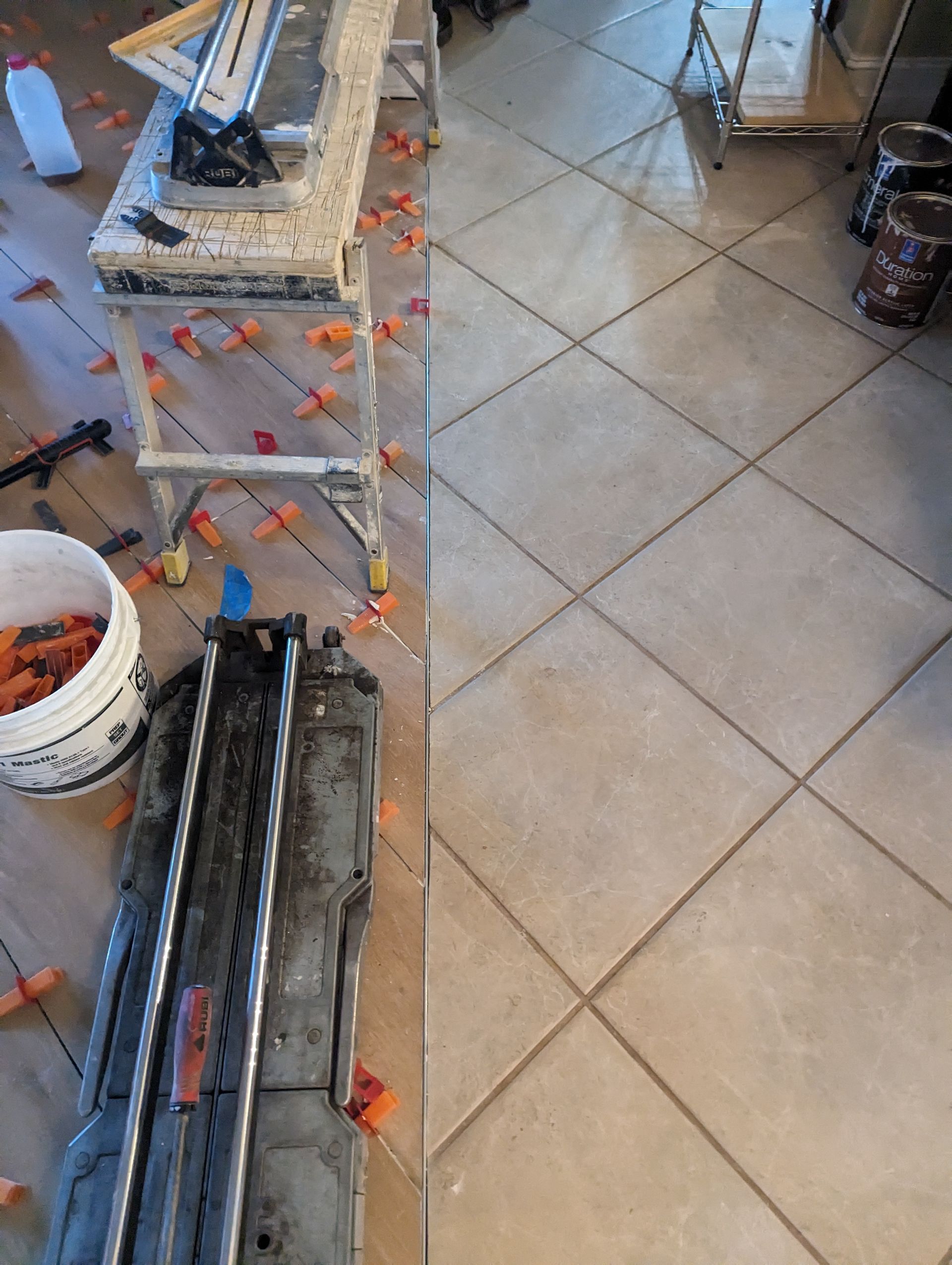 A tile cutter is sitting on a tiled floor next to a bucket of tiles.