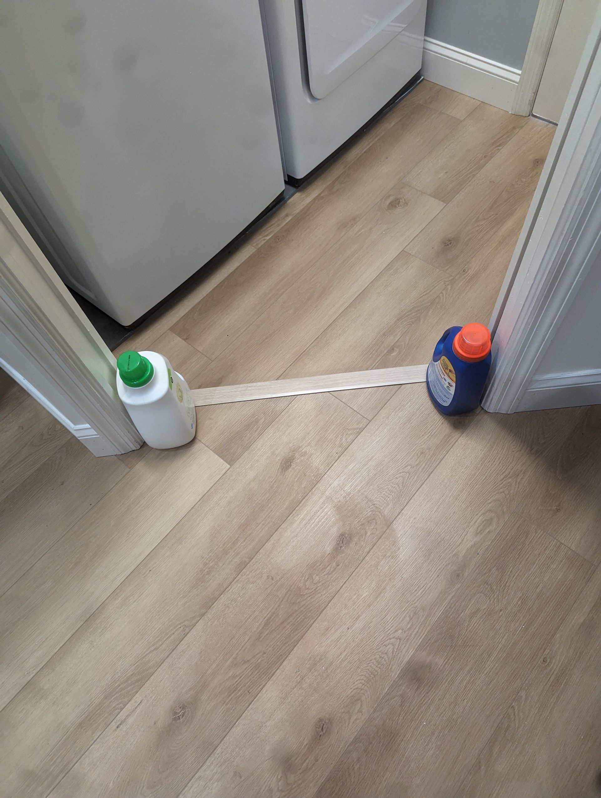 Two bottles of laundry detergent are sitting on a wooden floor in a laundry room.