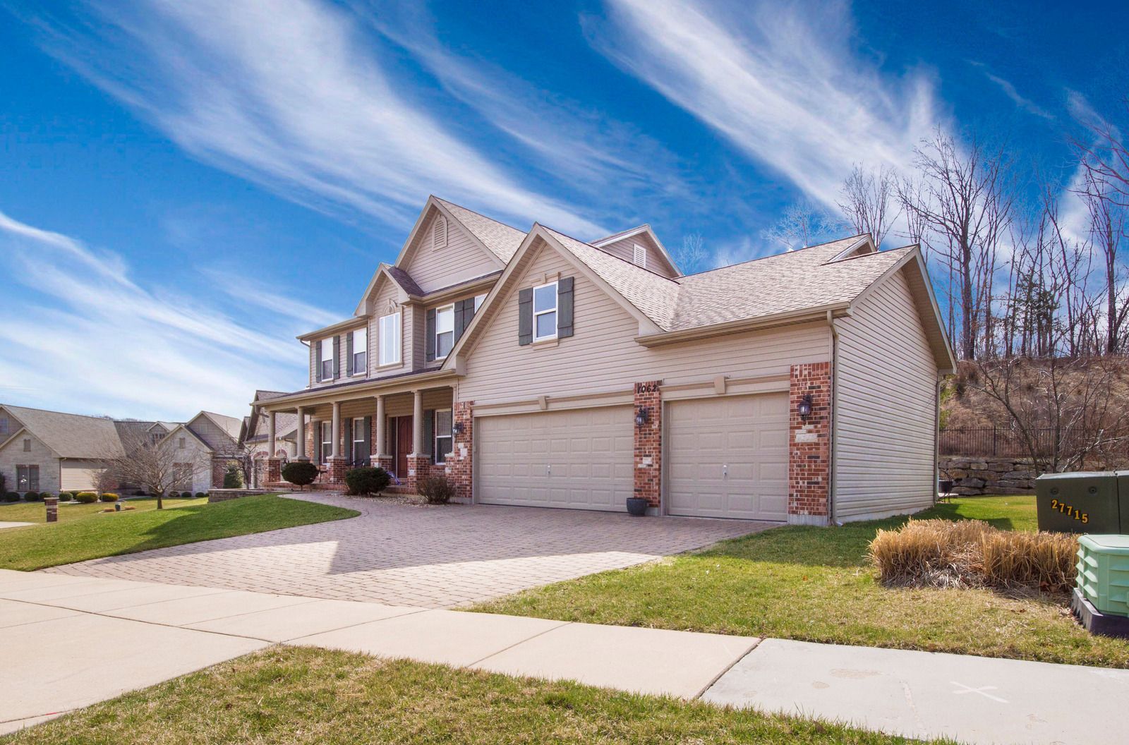 Two-story beige house with a two-car garage, brick accents, and a paved driveway under a blue sky with streaky clouds.
