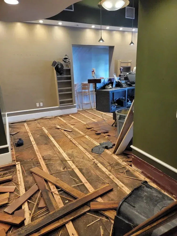 Room undergoing renovation; hardwood floor ripped up. Beige and green walls, with shelving unit.
